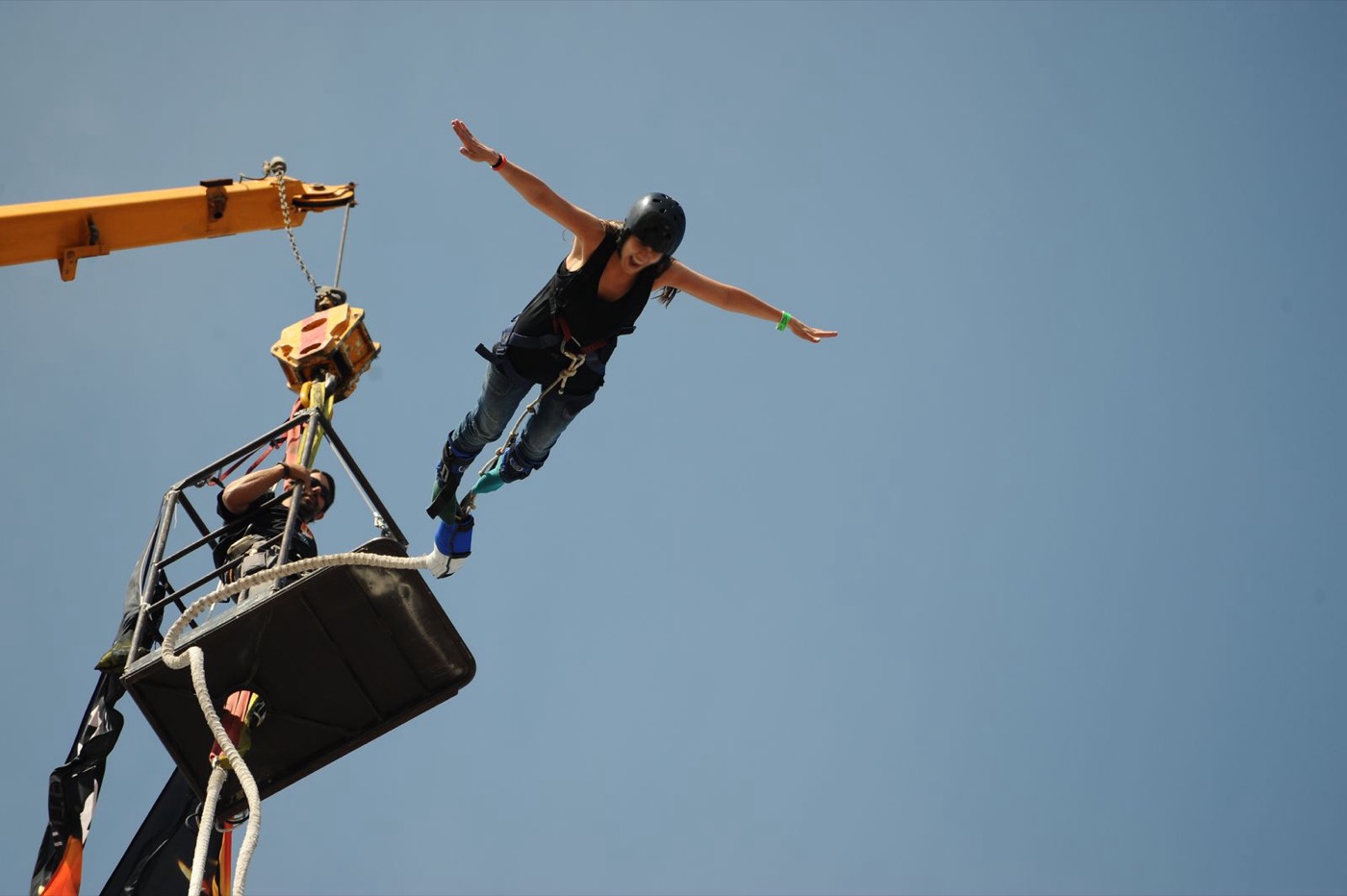 Jumper in a helmet and full harness leaping with arms spread while the crew watches from the platform