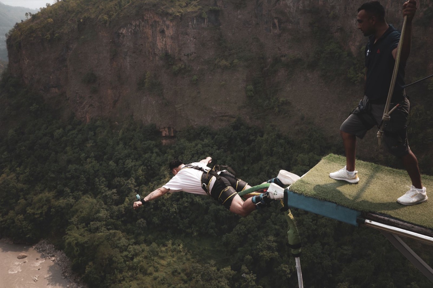First-time jumper diving off a bungee platform over a river canyon while the jumpmaster looks on